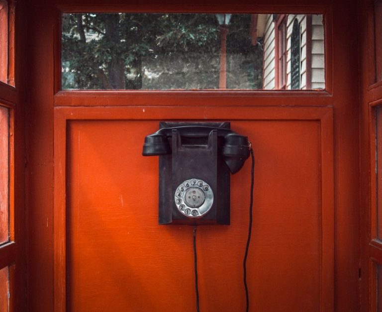 Schwarzes Wählscheibentelefon an oranger Wand, mit dem Hörer daneben.